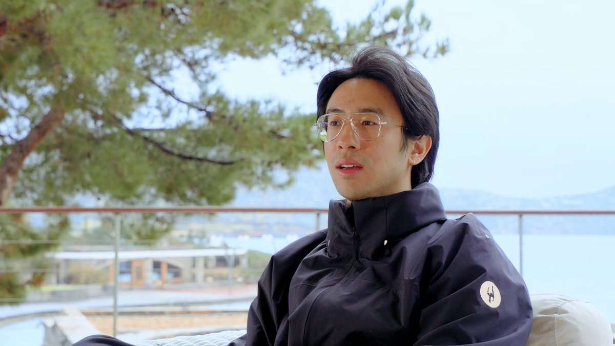 The image features a man wearing glasses sitting on a couch, looking off into the distance with his back to the camera. The background includes trees and water, creating an outdoor setting. There are also some objects like a keyboard and a laptop visible near the couch where the man is seated. The scene seems to be related to the post about tokenisation in finance or real-world assets, as it highlights the importance of access to real-world assets for blockchain technology.