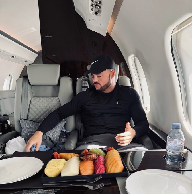 A man in athletic wear and a black cap sitting inside a private jet, looking down at a large bento-style meal containing salmon, pineapple, strawberries, and other foods.