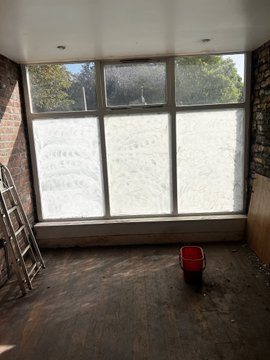 An empty room under renovation with a dirt floor, large windows, a red bucket in the center, and a metal ladder leaning against the left wall.