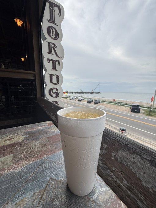A white styrofoam cup of coffee sitting on a wooden counter/bar surface, with a view through a window showing 'PORTUG' (Portugal) sign, a road with cars, and the ocean/sea in the background under a cloudy sky.