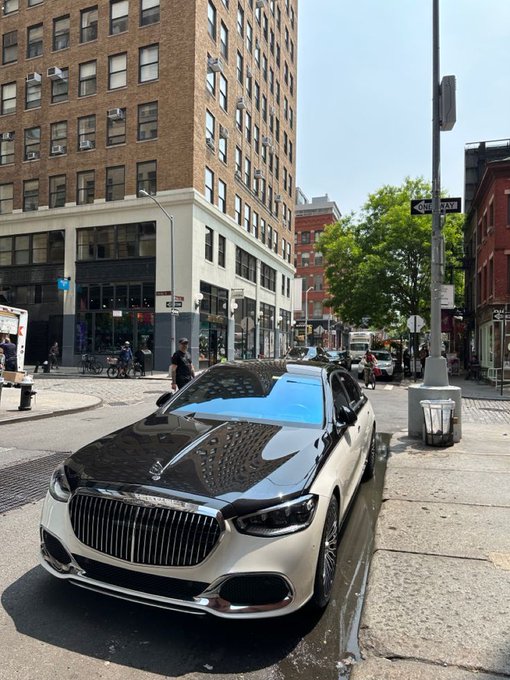 A black Mercedes-Benz C-Class car is parked on a city street, facing away from the camera. The car has a blue tinted windshield that reflects the surrounding environment. In the background, there are tall buildings lining the street, creating an urban atmosphere. A person can be seen walking nearby, and another individual is riding a bicycle further down the road.