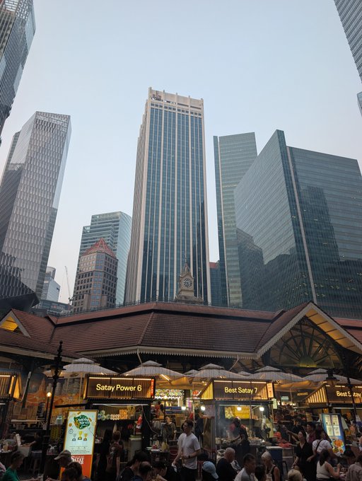 A bustling outdoor food market with stalls labeled 'Satay Power 6' and 'Best Satay 7' set against a backdrop of towering modern skyscrapers.