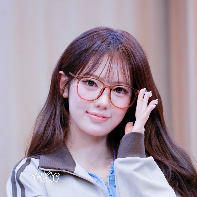 A young woman with long brown hair and bangs wears orange glasses and a white track jacket, gently touching her face with her manicured hand.