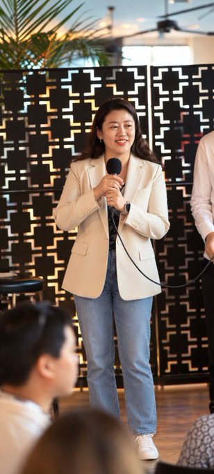 A young woman in a cream blazer and jeans holding a microphone while smiling, standing in front of a geometric black screen with seated audience members in the foreground.