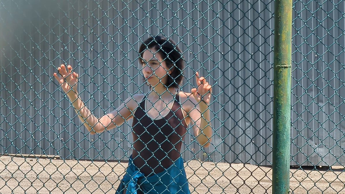 The image features a woman standing behind a chain-link fence, with her hands outstretched as if she's about to reach for something or someone outside of the frame. The photo has been edited to include text that reads "gonna watch my first WC because I clicked a few buttons on @jup_predict now every match is a season finale". This suggests that the woman might be participating in an online game, possibly related to sports or fitness, and she's excited about her performance.
