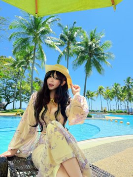 The image features a woman sitting on a bench by a pool, wearing a yellow dress with floral patterns. She has her hair styled in loose waves and is holding a hat over her head for shade. The background includes palm trees and an umbrella providing shade near the pool area. There are also other people present at the location but they are not the main focus of the image.