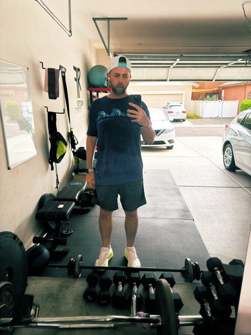 A man taking a mirror selfie in a garage gym, wearing a navy t-shirt, black shorts, pink sneakers, and a white cap backwards, surrounded by dumbbells and exercise equipment.