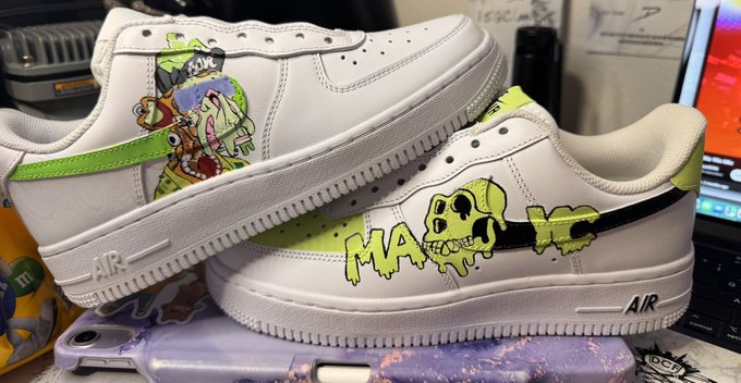 A pair of white Nike Air Force 1 sneakers featuring custom cartoon artwork in green and other colors, including characters resembling Nickelodeon cartoon figures, displayed on what appears to be a desk with a Nintendo device underneath.