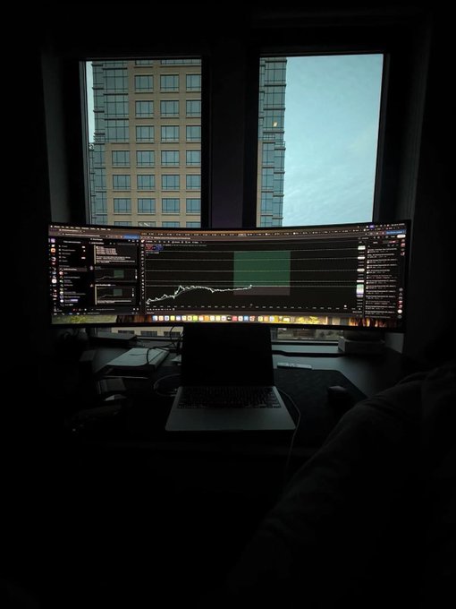 A dimly lit desk setup featuring a curved monitor displaying what appears to be a stock trading chart with green and red indicators, alongside a laptop and coffee cup, with city buildings visible through the window behind. The chart and trading interface visually reinforce the post's claim of making $5k from day trading by clicking buttons on such platforms.