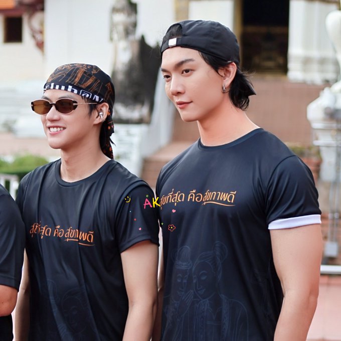 Two young men wearing matching black t-shirts with Thai text and bandanas stand together outdoors, appearing to be at a promotional event.