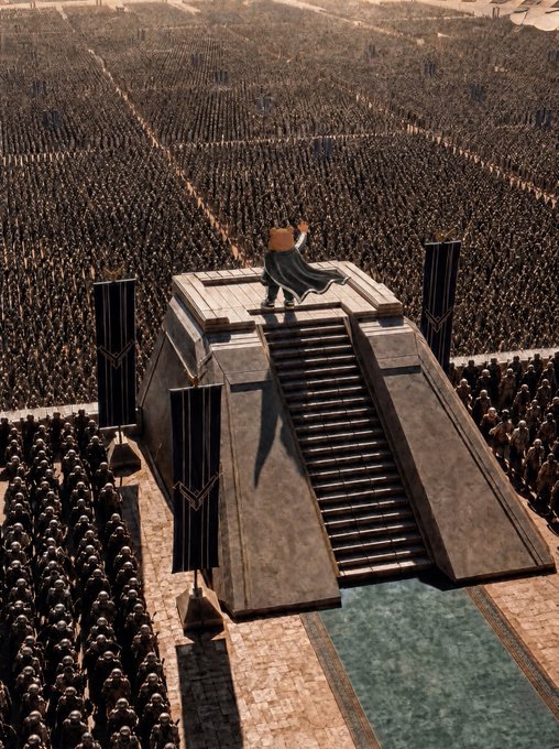 A figure in a dark cape stands atop a stone pyramid-like structure, raising one hand while addressing a massive army assembled in a vast desert landscape.