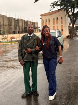 Two people wearing matching green and black blazers stand arm-in-arm outdoors on a wet pavement, smiling at the camera with a building and white vehicle in the background.