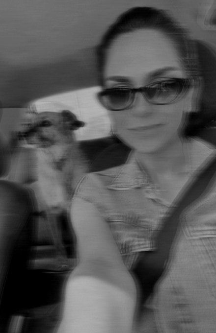 A blurry black-and-white selfie of a woman wearing sunglasses in a car with a dog beside her in the back seat.