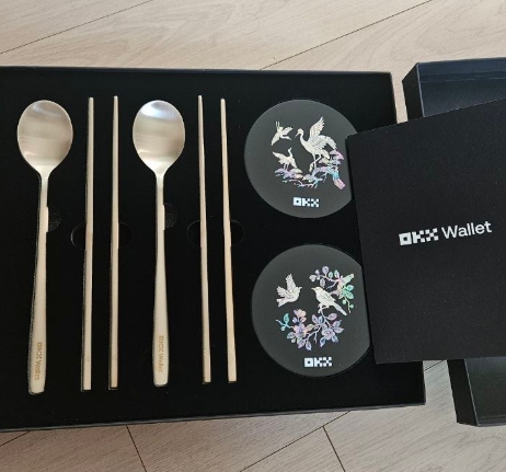 A Korean stainless steel cutlery gift set displayed in a black presentation box, including spoons and chopsticks, alongside two round containers and a 'OKX Wallet' branded box.