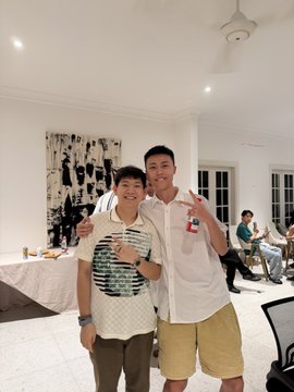 Two people pose together indoors, with the taller person wearing a white polo shirt and tan shorts, and the shorter person in a patterned vest.