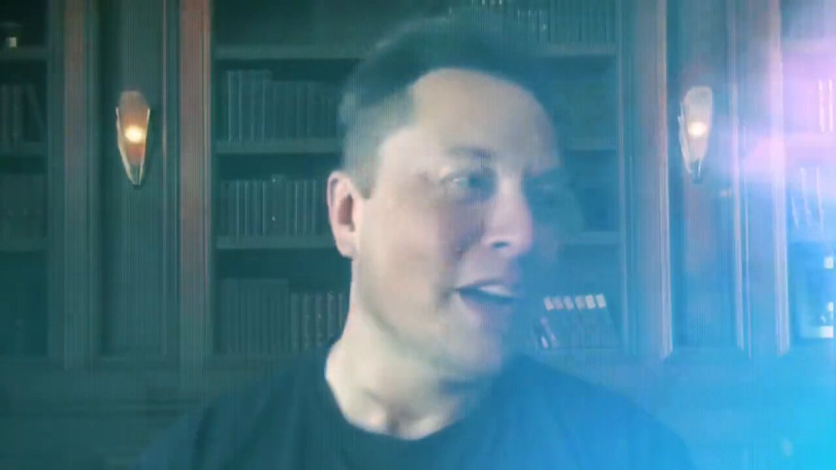 The image features a man with short hair wearing a black shirt, standing in front of a bookshelf filled with numerous books. The background includes two lights that seem to be glowing or emitting a blue light. There are also some books on display near these lights. The caption reads "Elon Musk thinks Dogecoin is pretty cool." This statement seems to be related to the man and his surroundings, as he appears to be in an intellectual environment surrounded by books.