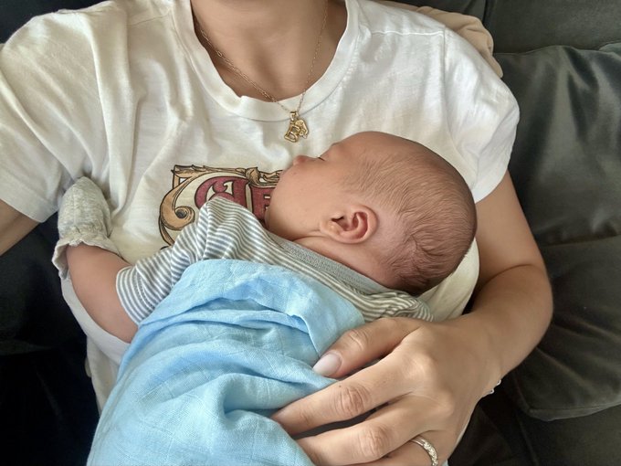 A person wearing a white t-shirt and a gold necklace holding a sleeping newborn baby wrapped in a striped blanket and blue cloth.