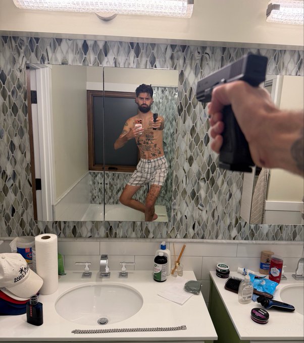 The image shows a man taking a selfie with his gun pointed at himself, while standing next to a bathroom sink. There are multiple bottles of various sizes placed around the sink area, suggesting that he might be preparing for or has just finished using the bathroom. The presence of these items and the man's action of pointing the gun towards himself indicate that this scene is likely related to a post about buying more soap at the store.

The image also contains text that reads "I bought more SOL here". This could possibly be an advertisement for a specific brand or product, such as soap, in relation to the bathroom setting and the man's actions.