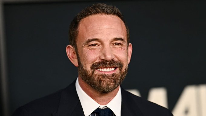 A headshot photograph of Ben Affleck smiling warmly while wearing a black suit, white shirt, and dark tie at what appears to be a formal event.