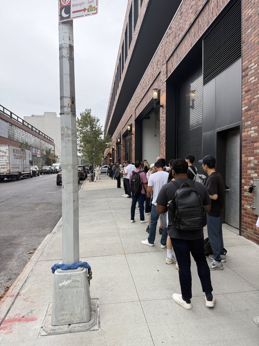 A line of people stands along a city sidewalk next to a brick building, waiting outside, with delivery trucks parked along the street to the left.