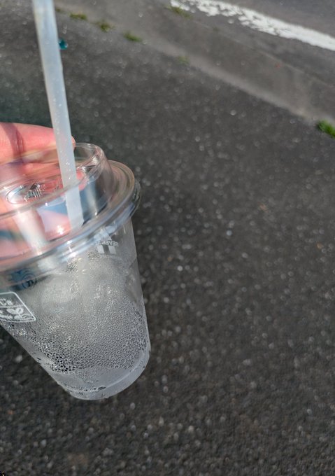 A close-up shot of a hand holding a clear plastic cup with a straw and condensation, set against a gray asphalt background.