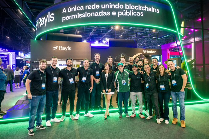 A group of people poses in front of a 'Rayls' booth at an event, with signage reading 'A única rede unindo blockchains permissionadas e públicas' and a 'tether' logo visible.