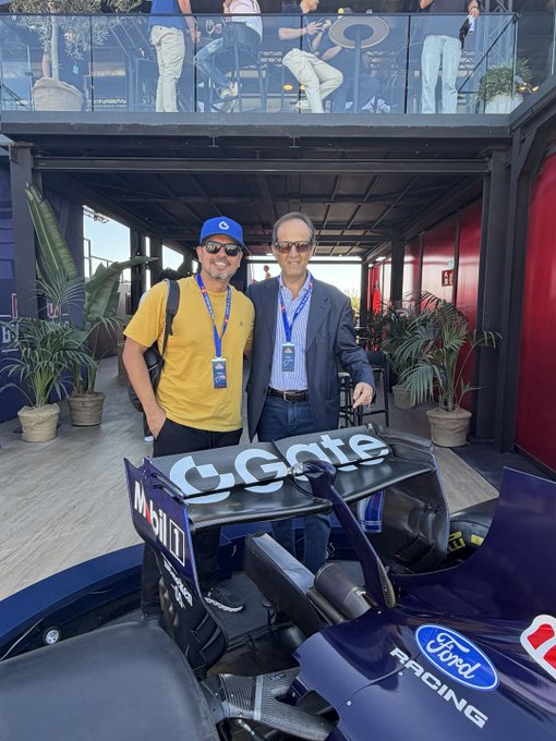 Two men posing together at a Formula 1 event, one wearing a yellow shirt and blue cap, standing next to a racing car with 'C.Gate' and 'Ford Racing' branding.