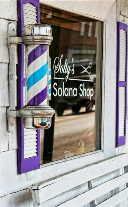 An exterior shot of a barbershop window with a vintage barber pole and white painted text on the glass reading 'Polly's Solana Shop' with scissors icon.