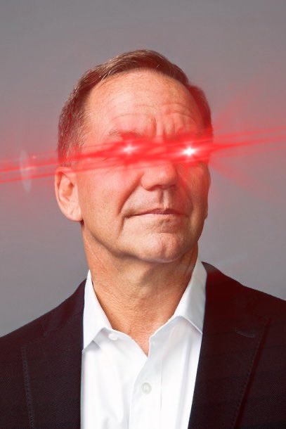 A portrait of a man in a white shirt and dark blazer with red laser beam effects edited across his eyes against a gray background.