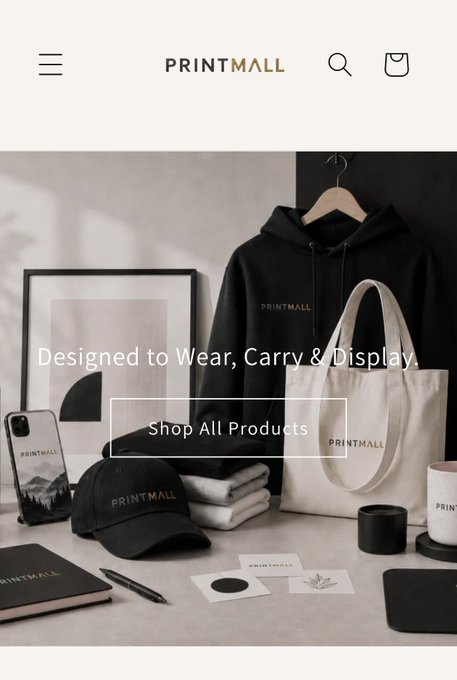 A homepage screenshot for PRINTMALL featuring the tagline 'Designed to Wear, Carry & Display' with a 'Shop All Products' button, showing branded merchandise including a black hoodie, tote bag, cap, phone case, mug, and stationery.