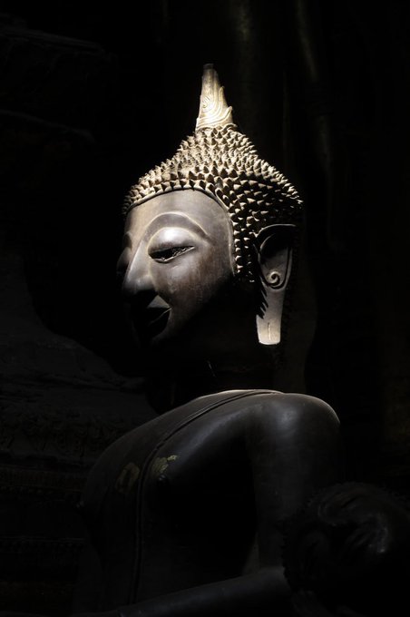 A dark, moody photograph of a Buddha statue, focusing on its serene face and intricate, pointed headpiece.