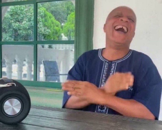 An elderly man in a dark traditional embroidered shirt laughing heartily at an outdoor wooden table, with a speaker visible on the left and a garden view through the window behind him.