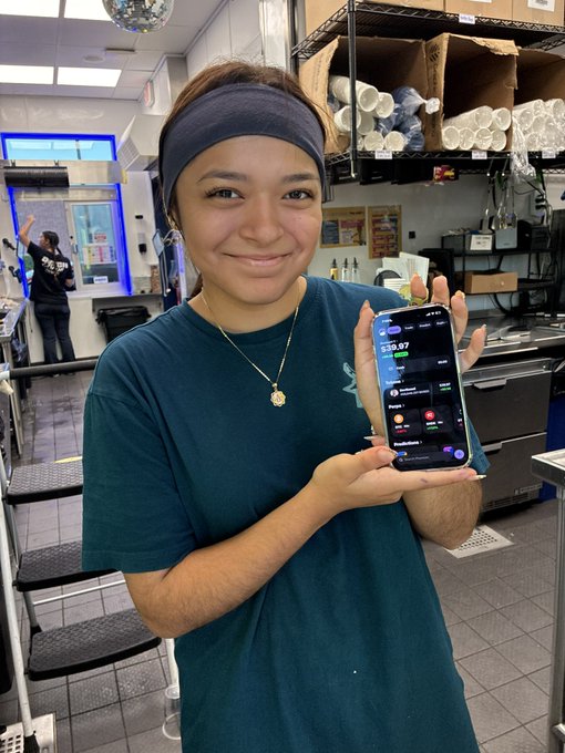 A young woman with a navy headband and teal shirt smiles while holding up a smartphone displaying a Robinhood-style investment app showing a $19.67 balance.
