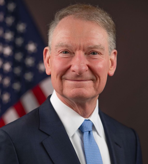An official portrait of an older man in a navy suit and light blue tie, smiling in front of an American flag.