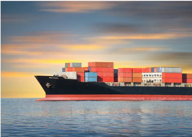 A large cargo ship loaded with colorful shipping containers (red, blue, and orange) sails across calm waters under a sunset sky with streaked clouds in orange and purple hues.