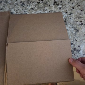 A person's hand holds open the blank, brown cardboard flap of a small, empty shipping box over a speckled granite countertop.