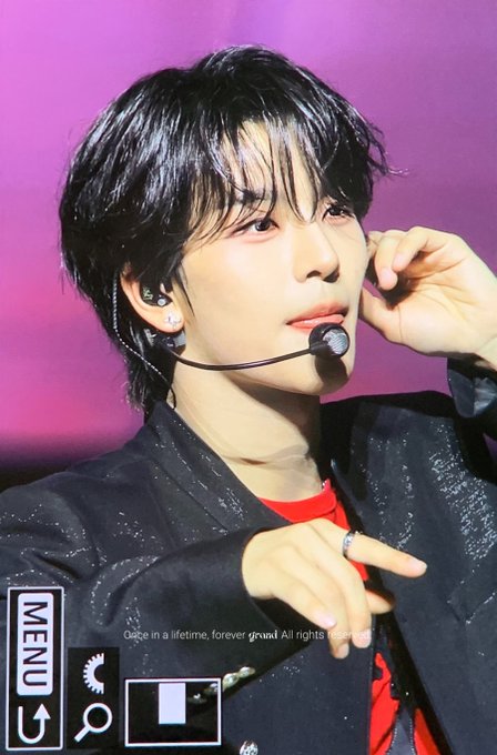The image features a young man wearing a black jacket, with his hair styled in a short cut. He has a microphone attached to his face, suggesting that he might be involved in some form of performance or live event. The background of the image is purple, which adds a vibrant and eye-catching element to the scene. There are also several other elements present in the image, such as a menu sign located at the bottom left corner and an arrow pointing upwards.