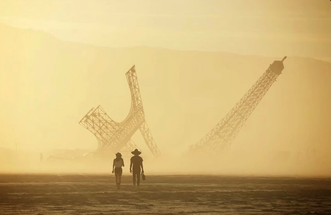 An atmospheric, hazy photograph of two silhouetted figures walking across a desert landscape toward a large, abstract sculptural structure resembling a hammer and sickle, with mountains barely visible in the dusty golden light.