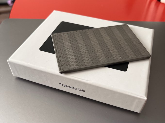 A gray rectangular electronic device with a grid pattern rests on its white packaging box labeled "Cryptotag Loki" on a gray table.