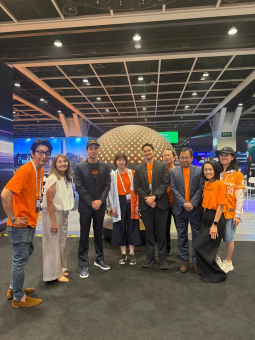The Metaplanet team poses together at Bitcoin Asia, with several members wearing orange shirts (one displaying "50"), standing in front of a large illuminated spherical installation on the main stage. The group, dressed in a mix of orange Metaplanet attire and business attire, smiles for the photo at the conference venue.