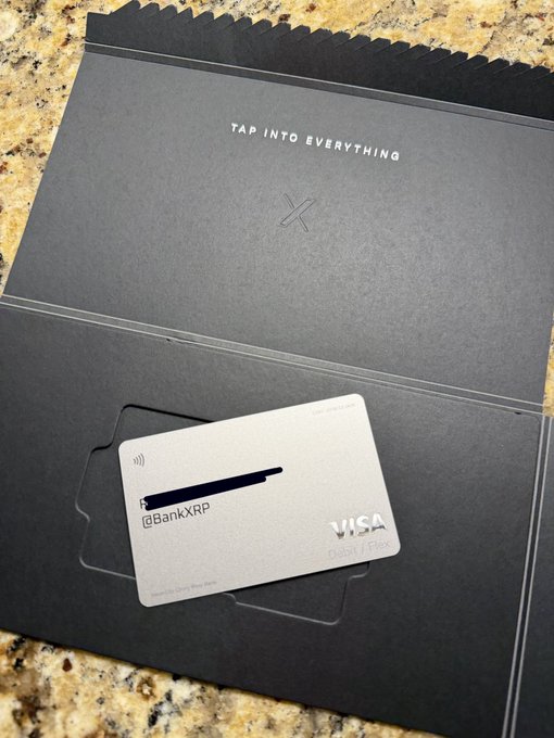 A new metal Visa debit card branded with "@BankXrp" and the X logo sits in its black envelope inscribed "TAP INTO EVERYTHING," unveiling the long-awaited X Money account. The card's sleek metallic finish reflects the premium, all-in-one financial features announced in the post—including 3% cashback, 6% APY, and fee-free global transactions.