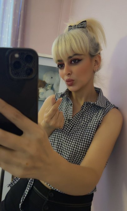 A blonde woman with a hair bow takes a mirror selfie in a black and white checkered top, puckering her lips at the camera while holding a black smartphone.