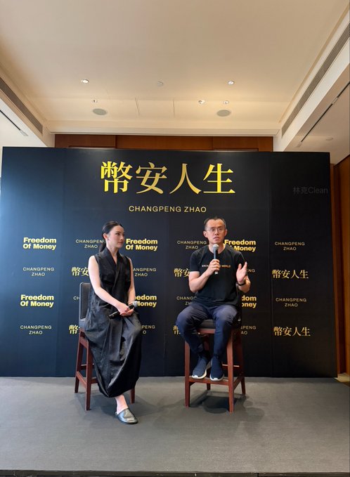 Photograph of a woman and a man seated on stage in front of a black backdrop with 'CHANGPENG ZHAO' and 'Freedom Of Money' text in both English and Chinese.