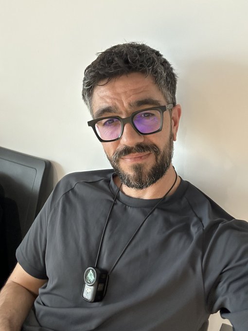 A selfie of a bearded man wearing glasses with purple-tinted lenses, a dark t-shirt, and a lanyard with a glowing pendant device.
