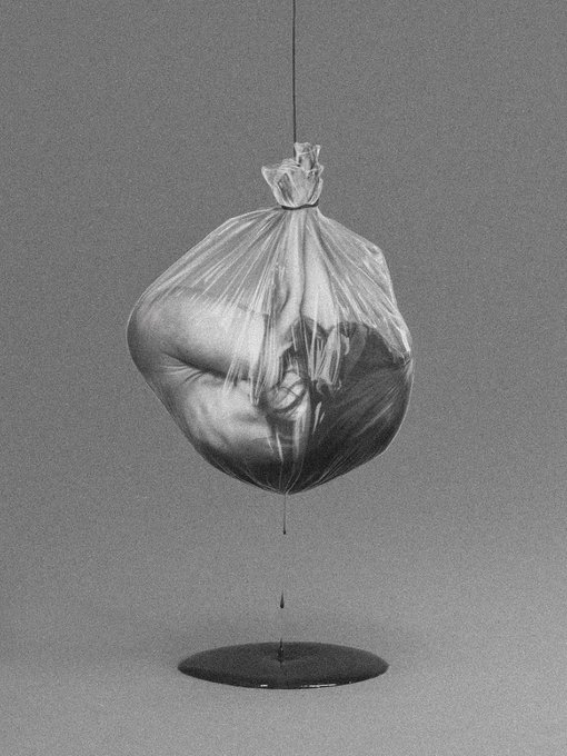 A surreal black-and-white image showing a transparent plastic bag suspended by a string, containing what appears to be a curled human figure, dripping a dark liquid onto a puddle below.