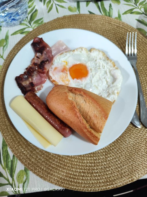 The image shows a plate of food, including eggs, bacon, cheese, bread, and sausage, placed on a table with a fork nearby. There are also two cups visible in the scene - one near the top left corner and another at the bottom right corner. The background features a green plant that adds a touch of nature to the setting. The post caption reads "#hive", which is likely related to the food being served on the plate, possibly indicating a social gathering or a shared meal among friends or family members.