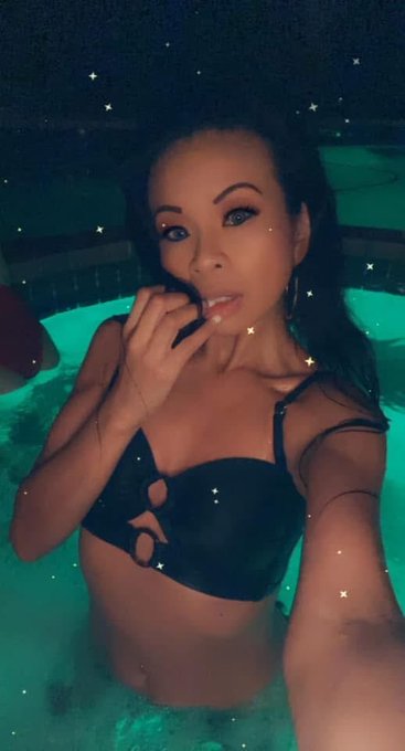 A woman in a black bikini poses in a swimming pool at night, with a sparkly filter overlay added to the selfie.