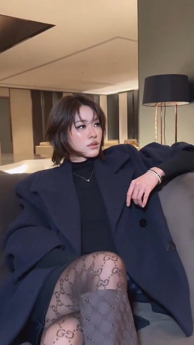 A woman with short brown hair poses on a gray couch wearing an oversized navy coat, black top, and patterned tights featuring the Gucci logo.