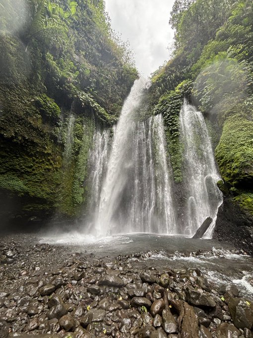 A tall waterfall cascades down lush, green moss-covered cliffs into a rocky stream bed below, with mist rising from the falling water.