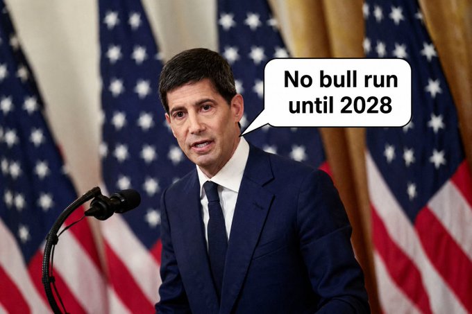 The image features a man dressed in a suit, standing at a podium with his mouth open as if he's speaking or giving a speech. The background of the image includes several American flags, which are arranged in rows behind the man. There is also an amusing text overlay on the image that reads "No bull run until 2028". This text seems to be a joke or a humorous commentary about the current political climate and the Fed's monetary policies.